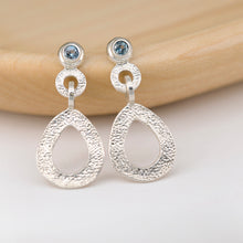 Load image into Gallery viewer, These hand-textured dangle post earrings features 3mm blue Montana sapphires. These earrings are positioned on a light stained wood. 