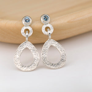 These hand-textured dangle post earrings features 3mm blue Montana sapphires. These earrings are positioned on a light stained wood. 