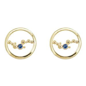 These stud earrings are handcrafted with solid 18K yellow gold, & a single 3mm Ceylon blue sapphire embraced in the middle of an abstract array of gold granulation. Against a white background. 