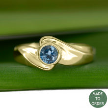 Load image into Gallery viewer, This ring, Timeless, embodies the elegance and harmony of the sea. This is a timeless solitaire ring with a unique flare. The ocean blue Montana sapphire is encircled by a graceful wave in 18K Yellow Gold. The band is finished with a high polish.