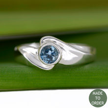 Load image into Gallery viewer, This ring, Timeless, embodies the elegance and harmony of the sea. This is a timeless solitaire ring with a unique flare. The ocean blue Montana sapphire is encircled by a graceful wave in Silver. The band is finished with a high polish.
